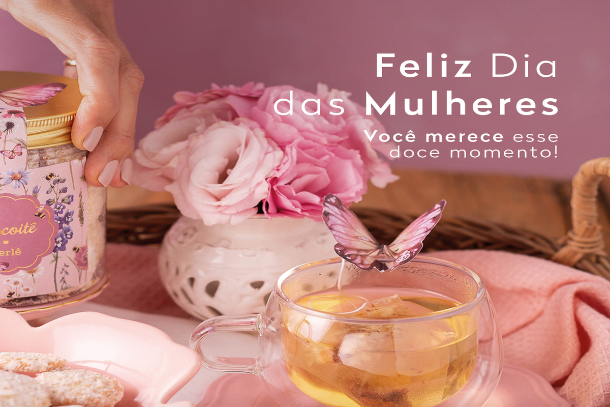 Mesa com chá servido em xícara de vidro, biscoitos polvilhados com açúcar e embalagem decorada da Biscoitê com borboleta na tampa. Ao fundo, flores rosas e cenário delicado em tons rosados celebrando o Dia das Mulheres.